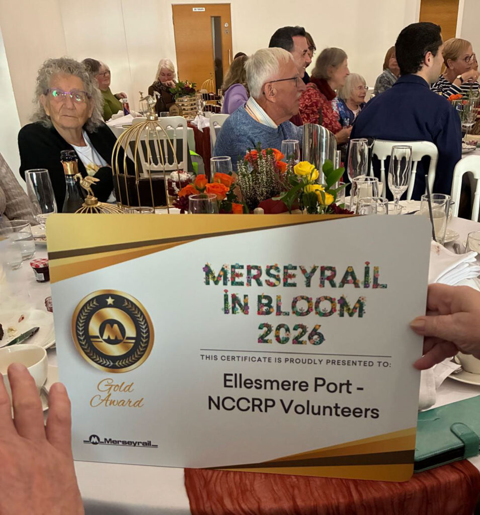 Merseyrail in Bloom 2026 Gold Award certificate for Ellesmere Port NCCRP volunteers, with Kenny and Carole Spain seated in the background.