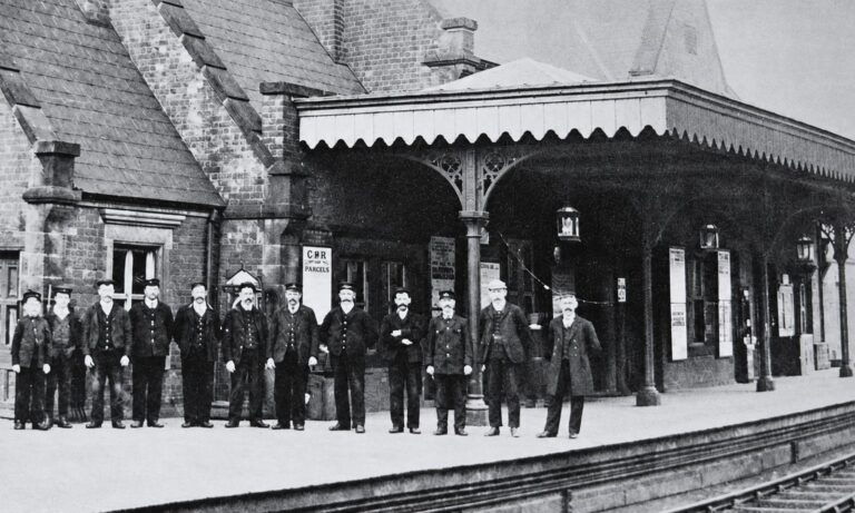 Frodsham Railway Station platform staff in the early 1900s, including the station master, ticket clerks, porters and young railway workers standing beneath the station canopy.
