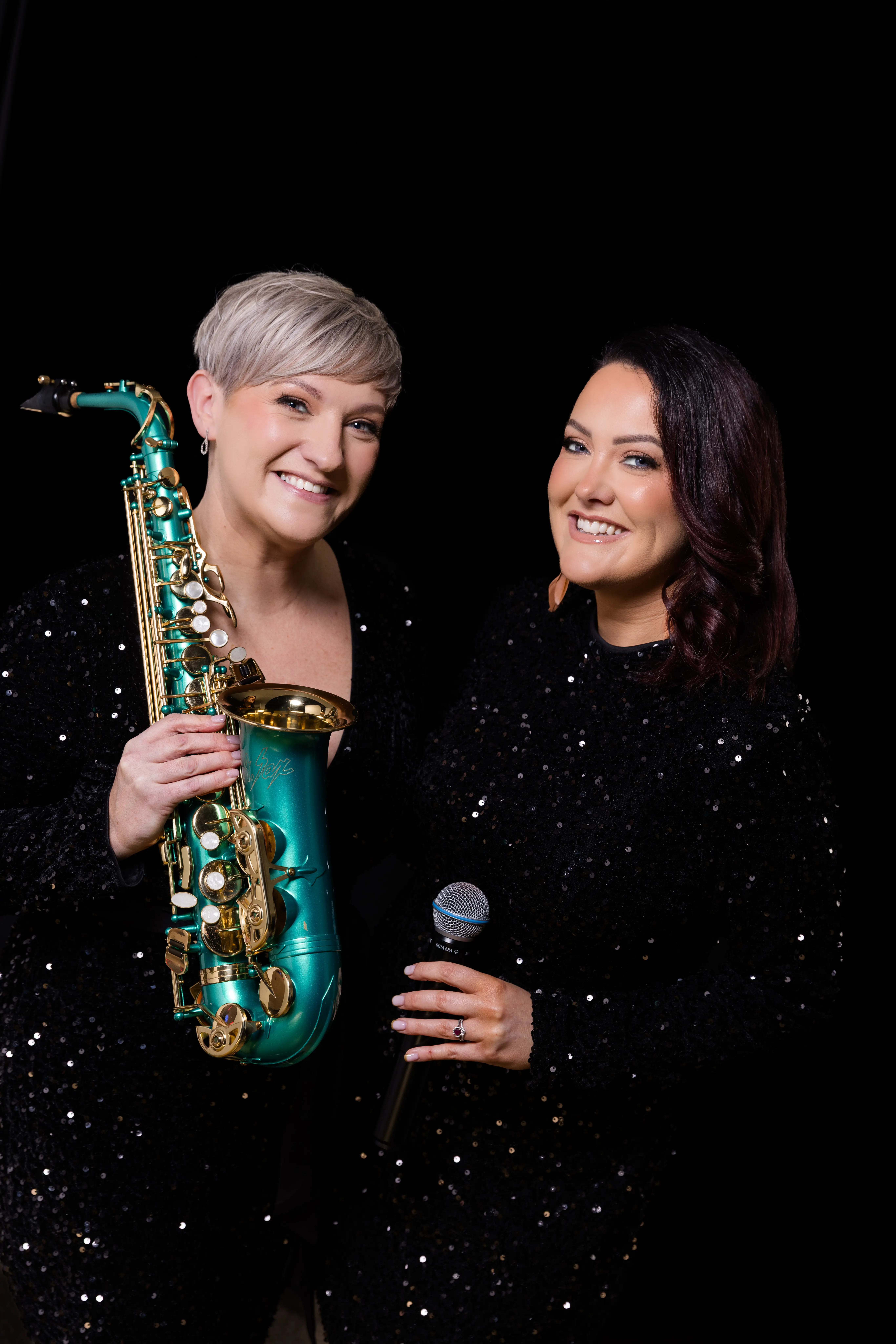 The Sax Pistols musical duo posing against a black background, with one member holding a turquoise saxophone and the other holding a microphone.